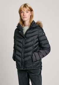 Zwarte pufferjack met chevron-quilting, voorzien van een met bont gevoerde capuchon, ritsluiting en zijzakken. Draagt over een lichtgroene top.