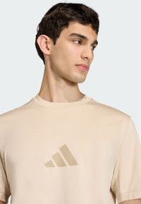 Camiseta de manga corta de color beige con una textura suave y un logo de Adidas en color claro en el pecho. Diseño simple con cuello redondo, ajuste casual.