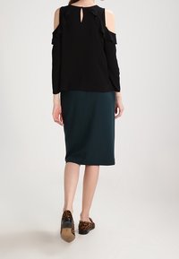 Svart långärmad topp med volangdetaljer och urtagna axlar, kombinerad med en mörkgrön knälång kjol och leopardmönstrade slip-on skor.