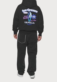 Felpa nera con cappuccio e grafica colorata "Ice Cream" sul retro, con testo in viola e blu, abbinata a jeans neri e sneakers bianche.