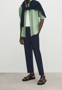 Ljusgrön linneskjorta, vit t-shirt, marinblå byxor och svarta lädersandaler. Tröja hängd över axlarna, minimalistisk design.