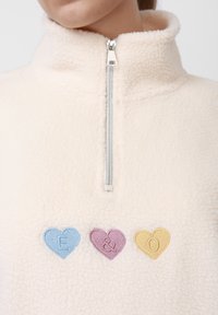 Sudadera de polar con media cremallera en color crema con superficie texturizada. Presenta tres parches con forma de corazón: "E" en azul, "&" en rosa y "O" en amarillo. Detalle de cremallera plateada.