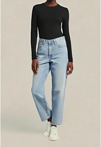 Personne portant un jean droit bleu clair, un haut noir à manches longues et des baskets blanches, debout devant un fond neutre.