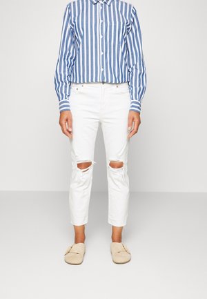 Personne portant une chemise à rayures verticales bleu et blanc à boutons, un jean blanc déchiré aux genoux, et des chaussures beiges sans lacets, debout devant un fond uni.