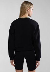 Sudadera negra recortada con hombros caídos, puños de canalé y dobladillo. Lleva puesta sobre pantalones cortos de ciclismo negros. Banda de cuello verde oliva visible en la parte posterior.