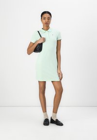 Pola kleita gaisā mentolzaļā krāsā, īsām piedurknēm, ar pogu apkakli, pielāgota forma, kopā ar melnu rokassomu un loaferiem, bežām ceļgalu augstuma zeķēm.