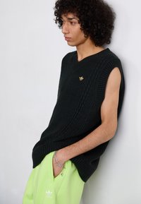 Pull sans manches en tricot noir avec un décolleté en V et un motif en câble. Associé à un pantalon vert fluo avec un détail de logo blanc.