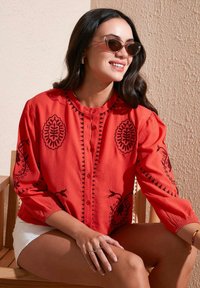 Rote, knopfbare Bluse mit schwarzen, gestickten Blumenmustern und einem strukturierten Stoff. Lange Ärmel mit elastischen Bündchen, runder Ausschnitt.