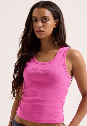 Frau mit langen braunen Haaren, die ein eng anliegendes rosa Tanktop mit der Aufschrift "Juicy" auf der Vorderseite trägt und vor einem schlichten Hintergrund posiert.