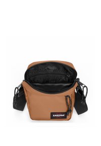 Brun tætsiddende crossbody taske med lynlåslukning, med sort indvendig og Eastpak-logo. Inkluderer justerbar rem og tekstureret overflade.