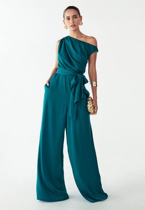 Turkooise jumpsuit met één schouder, wijde pijpen, een getailleerde taille en zijzakken. Heeft zachte, vloeiende stof en een bijpassende riem. Gouden accessoires.