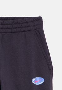 Calças de fato cinza escuro com cintura elástica, bolsos laterais e um emblema da Nike azul e rosa na coxa esquerda.