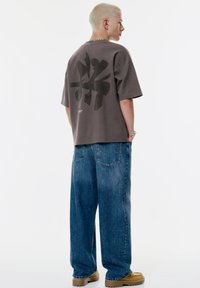 Grå oversized t-shirt med sort grafisk print på bagsiden, parret med lyse, løse jeans og tan-farvet fodtøj med en kraftig sål.