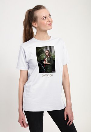 Woman with long ponytail wearing white tee showing "Gossip Girl" logo and photo of a woman in green plaid outfit holding red folder.