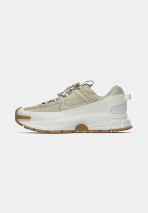Beige en witte sport sneaker met rubberen zool, elastische veters, gevoerde zool en gestructureerde hielpaneel, weergegeven in profiel op een witte achtergrond.