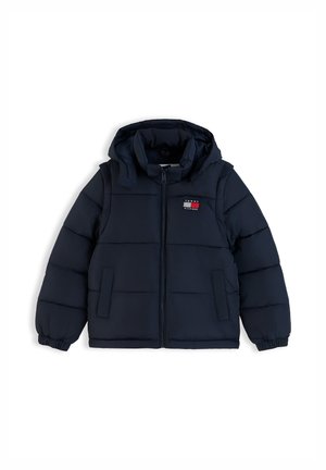 Giovane ragazza che indossa una giacca imbottita blu navy, camicia bianca, colletto a quadri rossi che sbuca, jeans a zampa di elefante e sneakers bianche, in piedi con le mani nelle tasche.