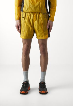 Gelbe, leichte Shorts mit elastischem Bund, kombiniert mit grauen Socken und schwarzen Laufschuhen mit orangefarbenen Akzenten.