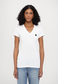 Camiseta blanca de cuello en V, hecha de algodón, de mangas cortas, con una textura suave, que presenta un pequeño parche de logo negro en el lado izquierdo del pecho.