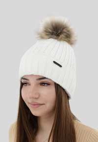 White knit beanie with textured ribbing and a brown fur pom-pom on top. Features a small black accent tag on the side.