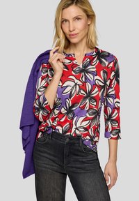 Mehrfarbige Bluse mit floralem Muster in Rot, Lila und Schwarz, V-Ausschnitt und 3/4-Ärmeln. Kombiniert mit dunklen Jeans und einem lilafarbenen Strickpullover.