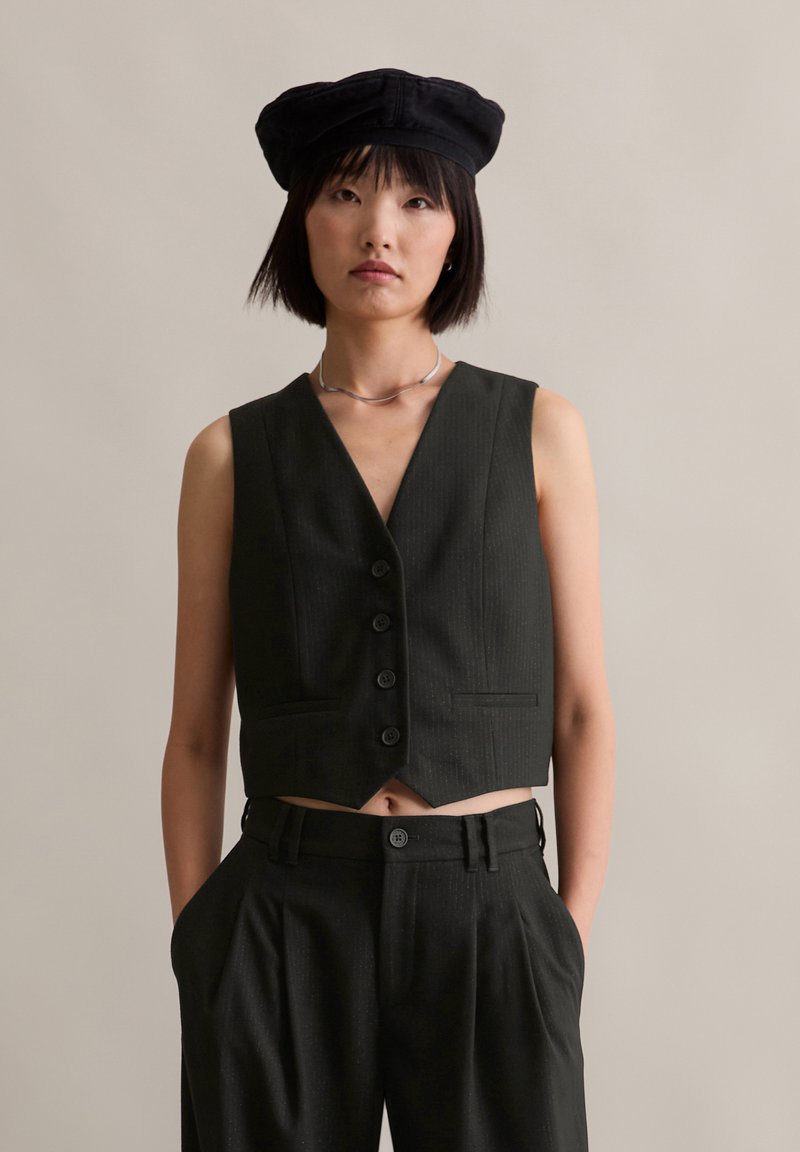 Schwarze, cropped Weste mit V-Ausschnitt, fünf Knöpfen und seitlichen Taschen, kombiniert mit hochgeschnittenen schwarzen Hose. Glatter, strukturierter Stoff.