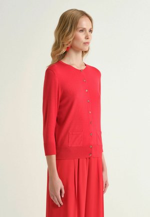 Donna con capelli biondi lunghi che indossa un cardigan rosso con bottoni e tasche e pantaloni ampi rossi coordinati, rivolta verso destra su uno sfondo chiaro.