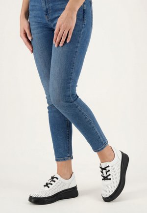 Person wearing fitted blue jeans and white perforated sneakers with black soles and laces, standing on a white floor.