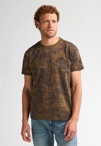 Bruine t-shirt met een gestructureerd landschapspatroon, ronde hals, korte mouwen en een getailleerde pasvorm, gedragen met een blauwe jeans.