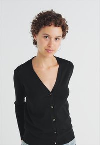 Cardigan negru cu decolteu în V, mâneci lungi și închidere cu nasturi. Fabricat dintr-un material moale, cu o textură netedă.