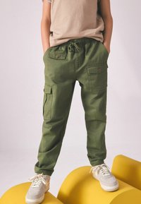 Grüne Cargo-Hosen mit elastischem Bund, Seitentaschen und Knopfdetails. Getragen mit einem hellbeigen T-Shirt und weißen Sneakers.