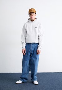 Grå hættetrøje med frontlomme og lille logo, parret med løse, lyseblå denimjeans og hvide sko. Brun kasket med emblem på fronten.