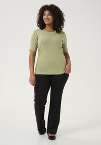 Světlezelený krátký rukávový top s kulatým výstřihem, v kombinaci s černými slim-fit kalhotami a černými plochými botami, na jednobarevném pozadí.
