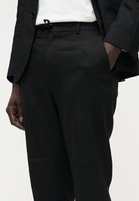 Άνδρας με μαύρο παντελόνι και σακάκι και άσπρο πουκάμισο, με το ένα χέρι στην τσέπη, εμφανιζόμενος από τη μέση έως το μισό μηρό.