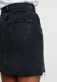 Jupe en denim noire avec ourlet effiloché, deux poches arrière et taille ajustée. La texture semble lisse avec des détails de couture subtils.