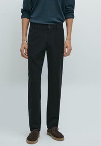 Schwarze maßgeschneiderte Hose mit geradem Beinschnitt. Aus glattem Stoff gefertigt, mit einem Knopfverschluss vorne und Taschen. Kombiniert mit braunen Schuhen.