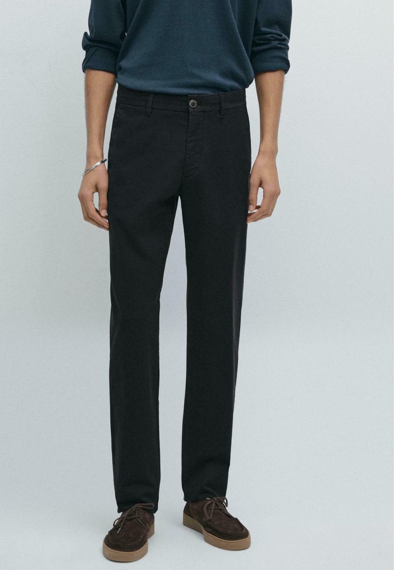 Schwarze maßgeschneiderte Hose mit geradem Beinschnitt. Aus glattem Stoff gefertigt, mit einem Knopfverschluss vorne und Taschen. Kombiniert mit braunen Schuhen.