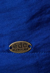 Blått tyg med en slät textur som har en metalltagg med guldtryck som säger "edc since 98." Twillväv med ett synligt sömnadsmönster.