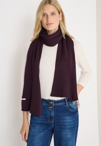 Geribbelde gebreide sjaal in dieppaars, om de nek gewikkeld, gecombineerd met een lichte trui en blauwe jeans, die een zachte textuur toont.