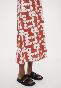 Röd midiklänning med ett geometriskt vitt mönster, kombinerad med svarta sandaler med öppen tå. Klänningen har en avslappnad passform och en mjuk textur.