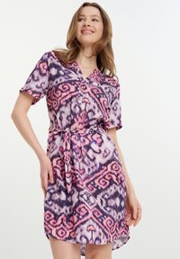 Kurzes Kleid mit kurzen Ärmeln, das ein lila, pinkes und weißes abstraktes Muster aufweist, einen V-Ausschnitt hat und an der Taille gebunden ist. Es besteht aus einem glatten Stoff.