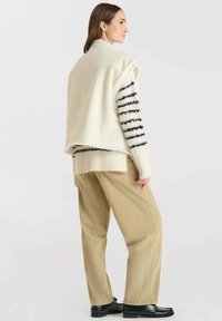 Ivore mouwloze vest over een zwart-wit gestreepte trui, gecombineerd met beige corduroy broek en zwarte schoenen. Gladde texturen en een relaxte pasvorm.