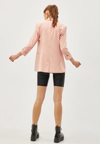 Rosa blazer med skräddarsydd passform, långa ärmar och bakre ventil, bärs över svarta cykelbyxor och kraftiga svarta stövlar. Slät textur.