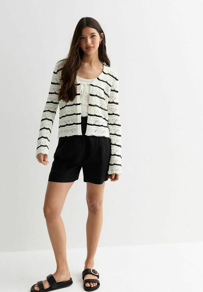 Weißer Strickcardigan mit schwarzen Streifen, mit langen Ärmeln und Spitzendetails, getragen über einem hellen Oberteil und schwarzen Shorts. Schwarze Sandalen.