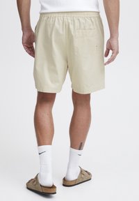 Shorts bej deschis, cu talie elastică, buzunar la spate și un material fin, ușor. Purtați cu șosete albe până la gleznă și sandale tip slip-on.