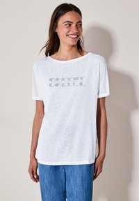 T-shirt blanc ample avec des manches courtes et des poignets retournés ; arbore le mot "BREEZE" en texte rayé vert sur le devant.