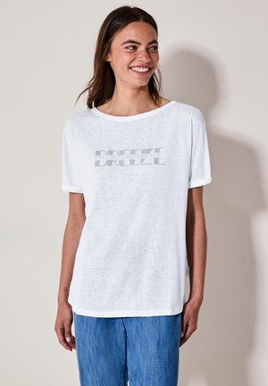 T-shirt blanc ample avec des manches courtes et des poignets retournés ; arbore le mot "BREEZE" en texte rayé vert sur le devant.