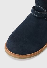 Marineblå semsket støvel med rund tå, sydd lys såle og en dekorativ stropp øverst. Glatt tekstur, minimale design detaljer.