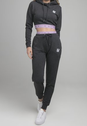 Kvinde iført mørkegrå cropped hoodie og matchende joggingbukser med lilla SikSilk taljebånd og hvide sneakers, der går fremad.
