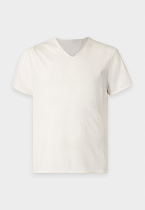 Homme aux cheveux noirs portant une chemise blanche à manches courtes, un pantalon marron ample et des baskets de couleur claire, debout devant un fond blanc uni.