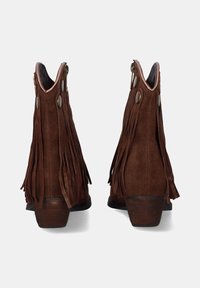 Botas de ante marrón hasta el tobillo con flecos largos, punta redonda y un tacón bajo estratificado. Presentan herrajes de latón y un diseño abierto en la parte superior.